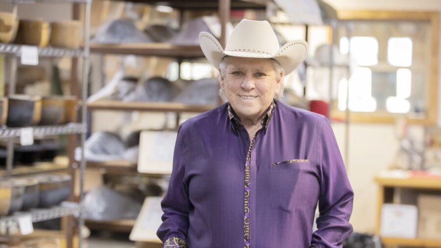 Lavonna ‘Shorty’ Koger stands and smiles for portrait in front of shelves of cowboy hats.